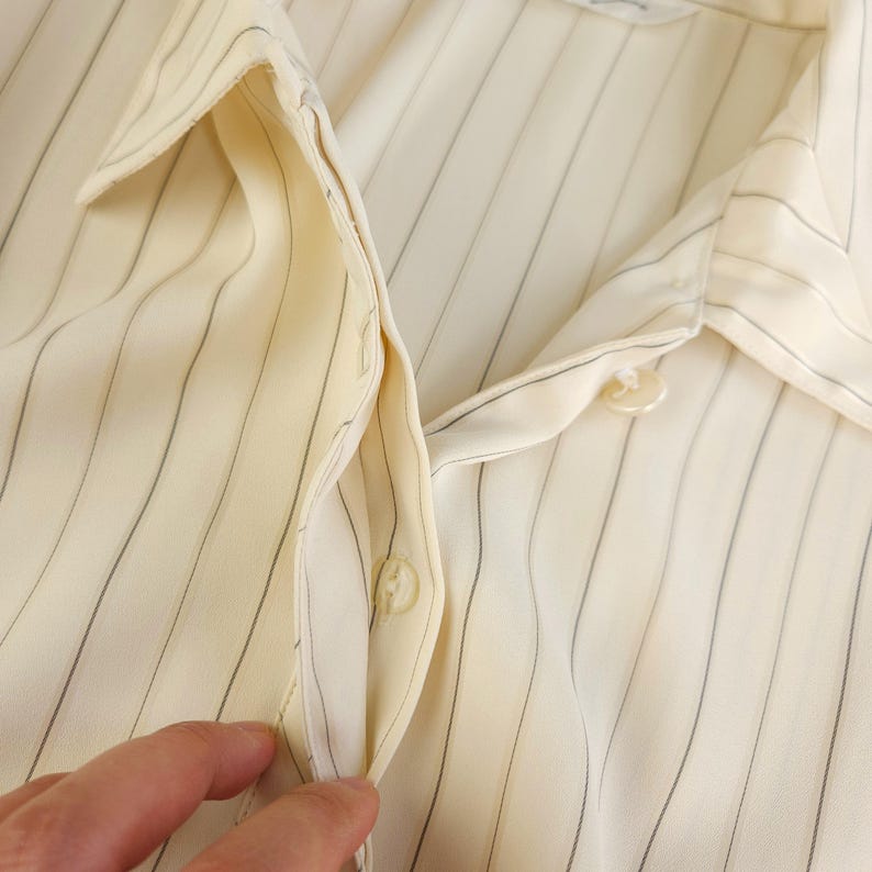 May include: Close-up of a cream-colored button-down shirt with thin vertical stripes. The shirt is unbuttoned at the collar and the top button. The fabric is smooth and silky.
