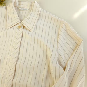 May include: A white striped long-sleeve shirt with a pointed collar and a button closure. The shirt has a subtle chevron design on the front.