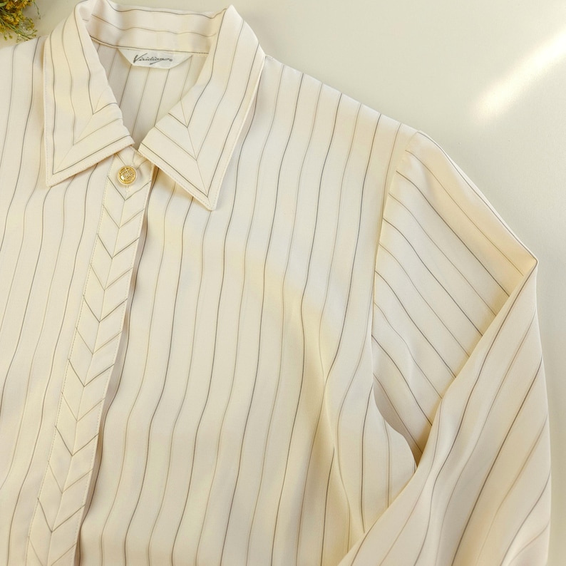 May include: A white striped long-sleeve shirt with a pointed collar and a button closure. The shirt has a subtle chevron design on the front.