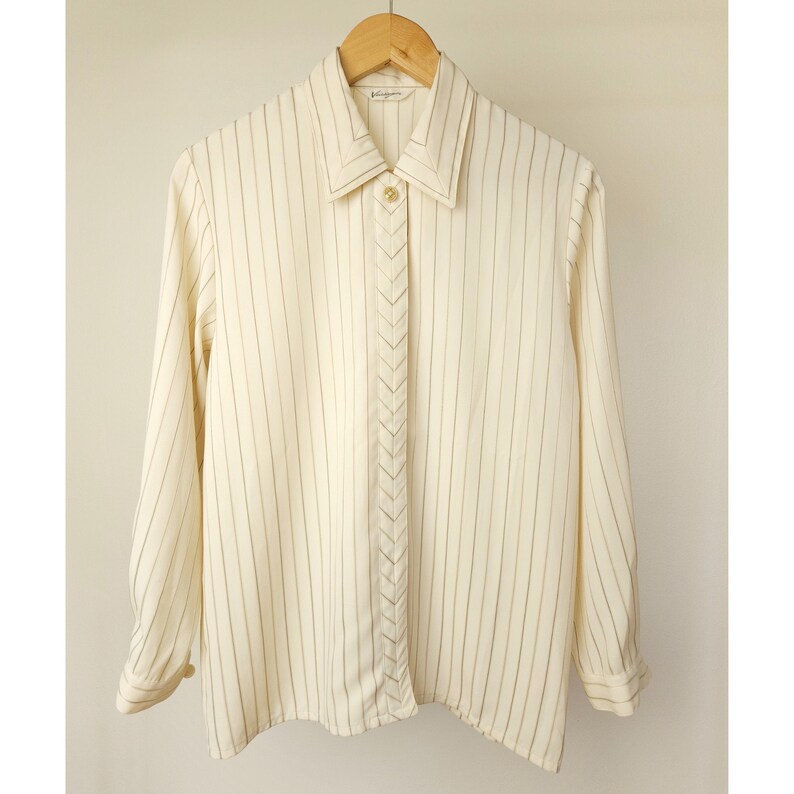 May include: A white long-sleeved button-down shirt with thin black stripes. The shirt has a pointed collar and a chevron-shaped detail down the front.
