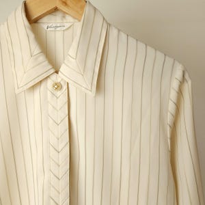 May include: A white button-down shirt with thin black vertical stripes. The shirt has a pointed collar and a single button closure. The label "Van Heusen" is visible on the collar.