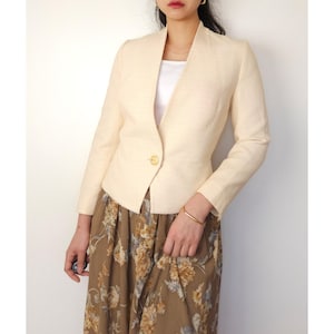 May include: A cream-coloured blazer with a single gold button and a wide collar. The blazer is fitted and has a slightly cropped length.