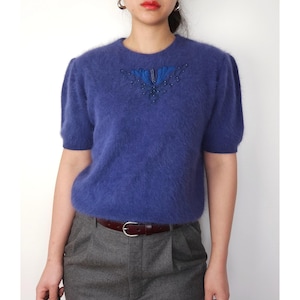 May include: A purple fuzzy sweater with a blue beaded embellishment on the front. The sweater has short sleeves and a round neckline.