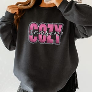 May include: Dark gray sweatshirt with the words "COZY season" in a pink and silver glitter design. The sweatshirt has a crew neck and long sleeves. The bottom of the sweatshirt has a fringe detail.