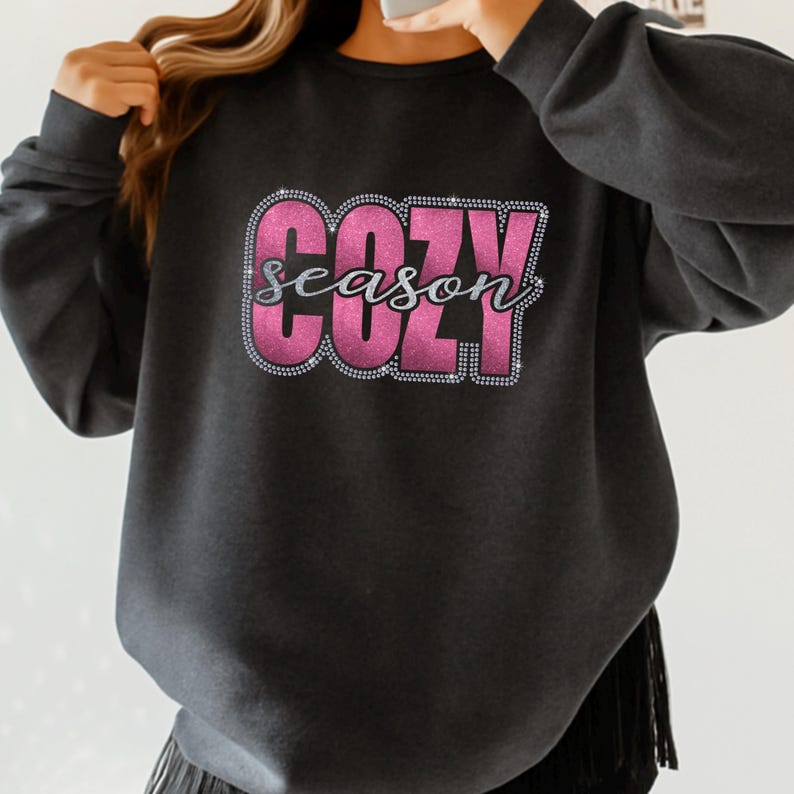 May include: Dark gray sweatshirt with the words "COZY season" in a pink and silver glitter design. The sweatshirt has a crew neck and long sleeves. The bottom of the sweatshirt has a fringe detail.
