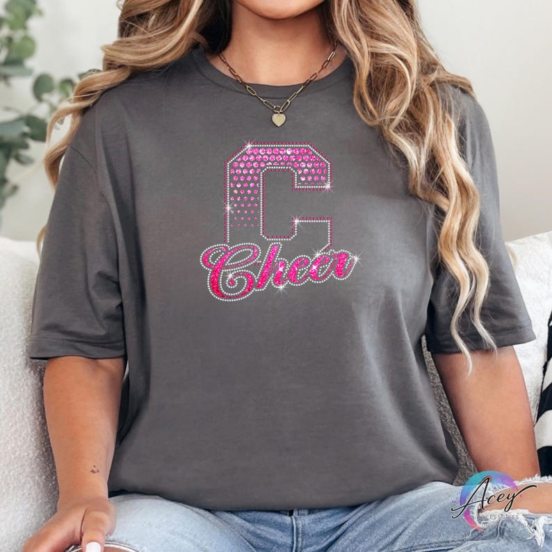 May include: A gray t-shirt featuring a large, pink rhinestone letter "C" with a heart pattern, and the word "Cheer" in a cursive font, also in pink rhinestones. The shirt is a casual style.