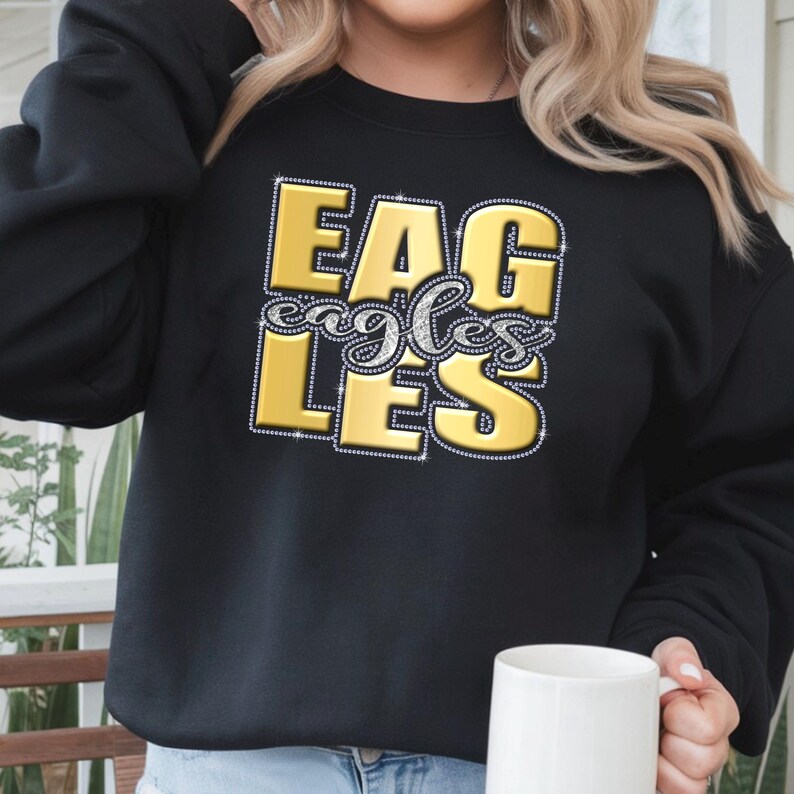 May include: Black sweatshirt with the word "EAGLES" in gold block letters outlined with rhinestones. The word "eagles" is written in silver cursive script. A white mug is held in the person's hand.