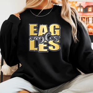 May include: Black sweatshirt with the word "EAGLES" in gold, outlined with rhinestones. The word "eagles" is written in a cursive font in the center of the design. The sweatshirt is a crew neck style.
