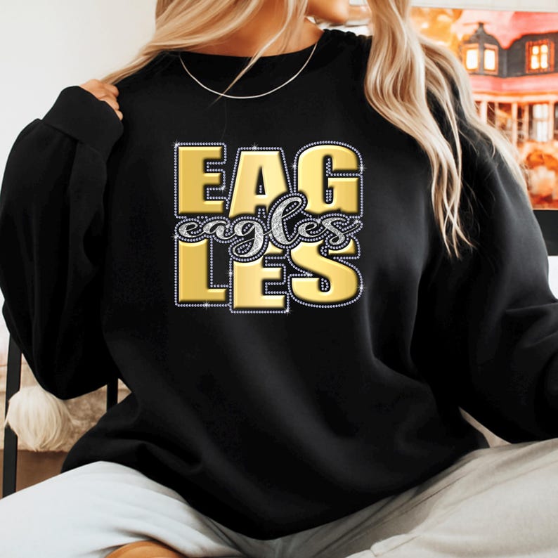 May include: Black sweatshirt with the word "EAGLES" in gold, outlined with rhinestones. The word "eagles" is written in a cursive font in the center of the design. The sweatshirt is a crew neck style.