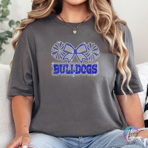 Pu&ograve; includere: Una t-shirt grigio antracite a maniche corte con un design grafico centrale realizzato in strass blu e argento. Il motivo mostra un grande fiocco affiancato da due pon-pon sopra la parola BULLDOGS in un carattere sans-serif grassetto.