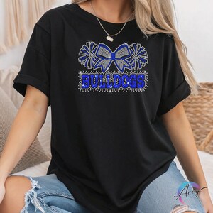 Pu&ograve; includere: Una t-shirt nera a maniche corte con un design grafico realizzato in strass blu e argento. Il motivo mostra un grande fiocco affiancato da due pon-pon sopra la parola BULLDOGS in un carattere stampatello in grassetto, tutto in pietre scintillanti.