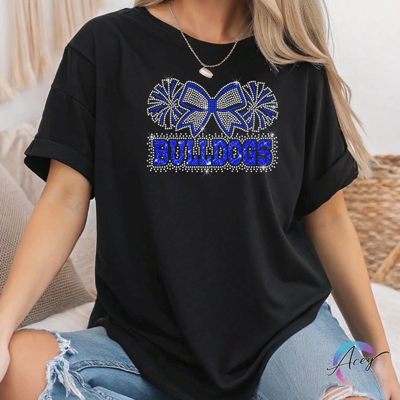 Pu&ograve; includere: Una t-shirt nera a maniche corte con un design grafico realizzato in strass blu e argento. Il motivo mostra un grande fiocco affiancato da due pon-pon sopra la parola BULLDOGS in un carattere stampatello in grassetto, tutto in pietre scintillanti.