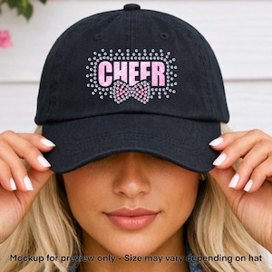 May include: Black baseball cap with the word "CHEER" in pink rhinestones, outlined with clear rhinestones. A pink bow is centered below the word. The cap is a classic, adjustable style.