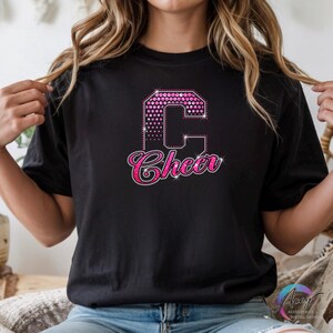 May include: Black t-shirt with a pink rhinestone design. The design features a large letter "C" and the word "Cheer" in a script font, both outlined in pink rhinestones. The shirt is a crew neck.