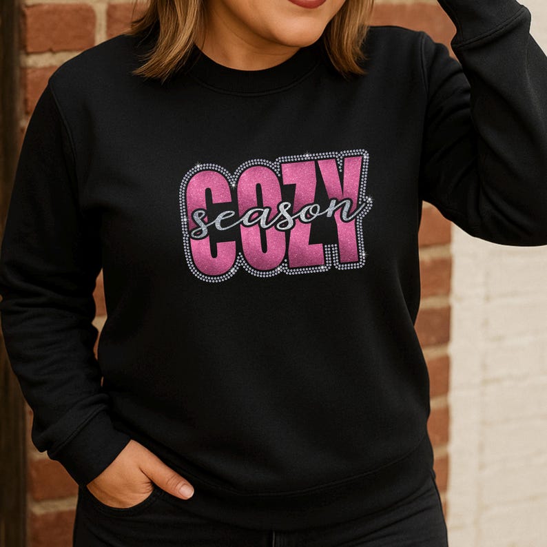 May include: Black sweatshirt with the words "COZY season" in a pink, rhinestone-outlined design. The word "COZY" is in a bold, block letter font, while "season" is in a cursive font. The sweatshirt is a crew neck style.