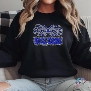 Pu&ograve; includere: Una felpa nera a maniche lunghe con un design grafico centrale realizzato in strass blu e argento. Il disegno raffigura un grande fiocco affiancato da due pon pon, con la parola BULLDOGS scritta in grassetto blu sotto il fiocco.