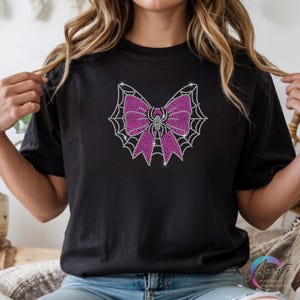 Puede incluir: Una camiseta negra de manga corta con un dise&ntilde;o gr&aacute;fico central hecho de peque&ntilde;os brillantes centelleantes. El dise&ntilde;o muestra un lazo rosa brillante sobre un patr&oacute;n de telara&ntilde;a blanca, con una peque&ntilde;a ara&ntilde;a en el centro del lazo.