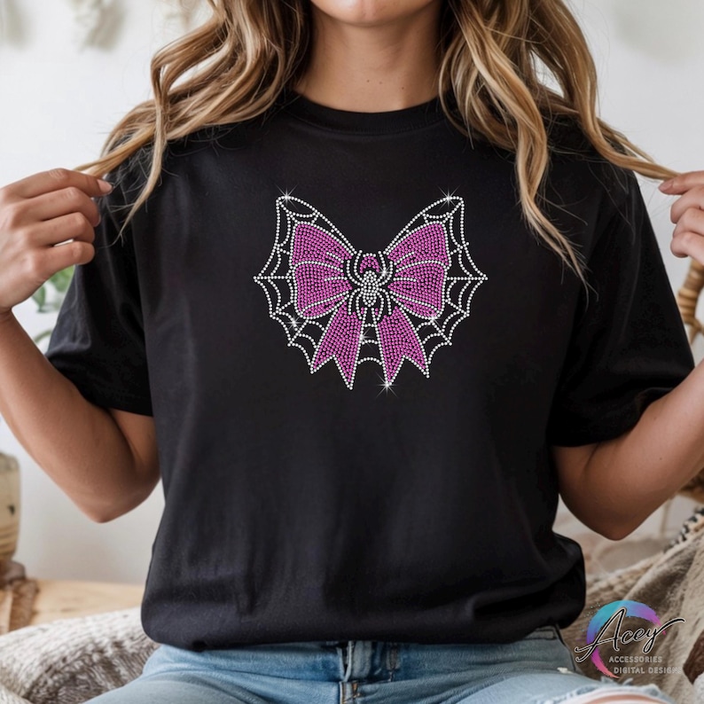 Puede incluir: Una camiseta negra de manga corta con un dise&ntilde;o gr&aacute;fico central hecho de peque&ntilde;os brillantes centelleantes. El dise&ntilde;o muestra un lazo rosa brillante sobre un patr&oacute;n de telara&ntilde;a blanca, con una peque&ntilde;a ara&ntilde;a en el centro del lazo.