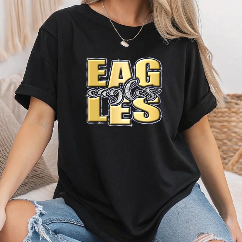 May include: Black t-shirt with the word "EAGLES" in gold block letters, with "eagles" in a script font. The letters are outlined with a silver glitter effect. The shirt is a casual style.