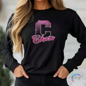May include: Black long-sleeve shirt with a large pink rhinestone letter "C" and the word "Cheer" in a script font. The letter "C" has a dotted pattern. The word "Cheer" is in a cursive font.