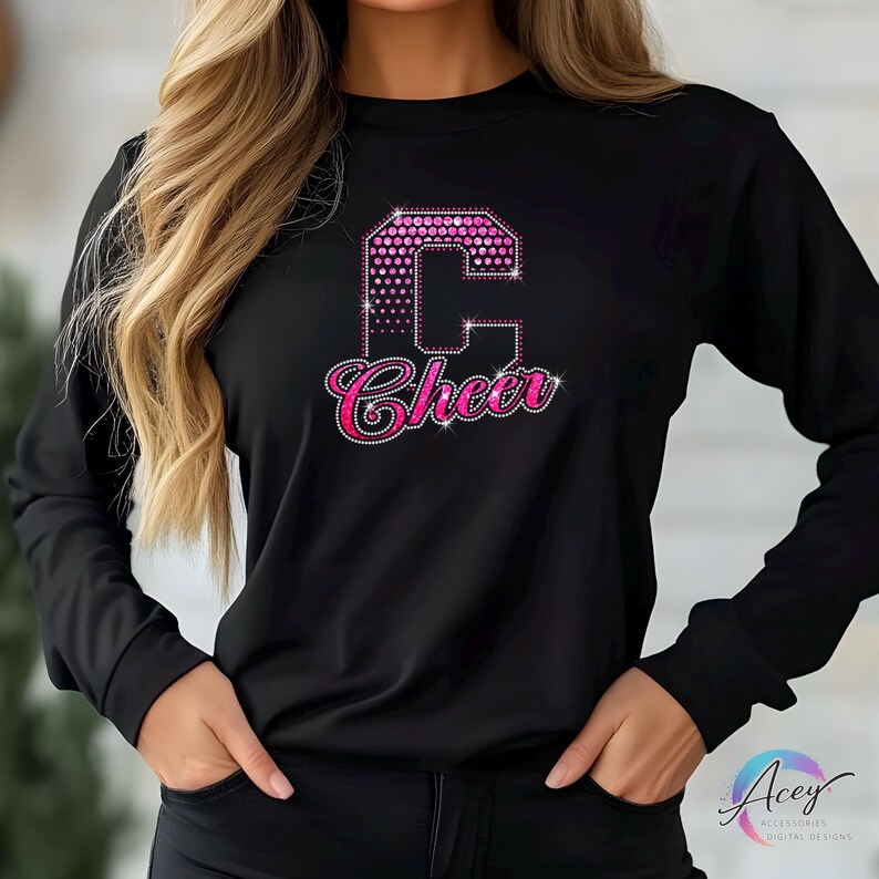 May include: Black long-sleeve shirt with a large pink rhinestone letter "C" and the word "Cheer" in a script font. The letter "C" has a dotted pattern. The word "Cheer" is in a cursive font.