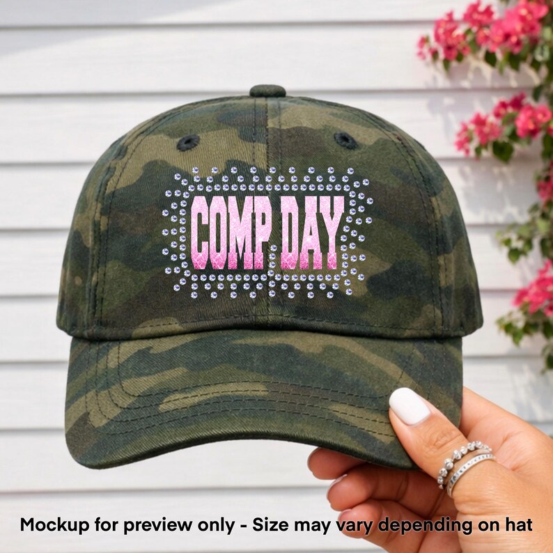 May include: Camouflage baseball cap with the text "COMP DAY" in pink, outlined with rhinestones. The hat has a curved brim and a button on top. The background is a white wooden wall with pink flowers.