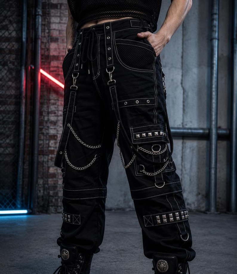 May include: Black cargo pants with white stitching, metal rivets, and chains. The pants feature multiple pockets, straps, and a drawstring waist. The design has a punk or alternative aesthetic.