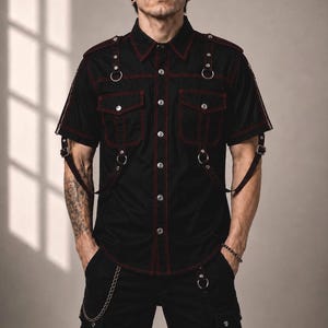 Peut inclure: Une chemise noire à manches courtes boutonnée avec des surpiqûres rouges contrastantes, deux poches de poitrine à rabat, des épaulettes et des sangles décoratives avec des anneaux en métal argenté et des boucles sur le devant et les manches.