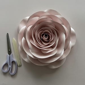 Puede incluir: Una gran rosa de papel rosa rubor con múltiples capas de pétalos. Unas tijeras con mangos morados claros y una plegadera están junto a la flor. La rosa es un suministro artesanal para hacer flores de papel.