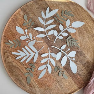 Könnte beinhalten: Ein Holzserviertablett mit natürlicher Holzmaserung, verziert mit ausgeschnittenen Blättern aus Papier in Hellblau und gedämpftem Grün. Die Blätter sind in einem dekorativen Muster angeordnet und schaffen ein botanisches Thema.