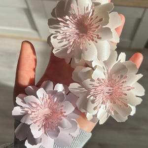 Könnte beinhalten: Drei zarte Papierblumen in Weiß- und Rosatönen. Die Blumen haben mehrere Schichten von Blütenblättern und ein detailliertes Zentrum. Die Blumen werden in einer Hand gehalten, was ihr kompliziertes Design und ihre sanfte Farbpalette hervorhebt.