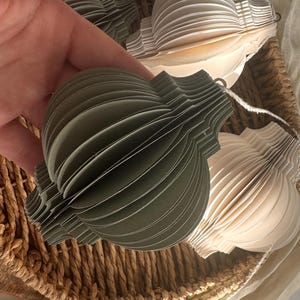 Può includere: Una mano tiene in mano un ornamento di carta verde con un design sferico a strati. Altri ornamenti di carta color crema e verde sono visibili in un cestino di vimini. Gli ornamenti sono infilati con un filo bianco, suggerendo un uso decorativo.
