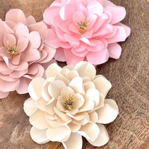 May include: Three artificial flowers in shades of pink and cream sit on a wooden surface. The flowers have multiple layers of petals and a golden centre. These flowers are a decorative item.