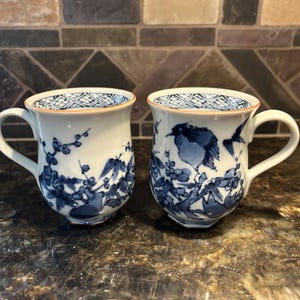 May include: Two white ceramic mugs with blue floral and bird designs. The mugs have a handle and a decorative rim. The interior rim features a geometric pattern. The mugs are on a dark surface.