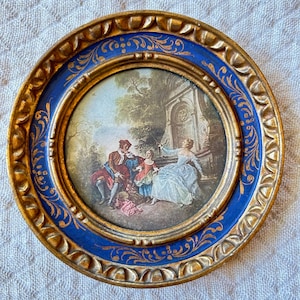 May include: A circular decorative plate featuring a central painting of three figures in a garden setting. The plate has a wide, deep blue border decorated with gold filigree patterns and a textured, scalloped gold outer rim edge.