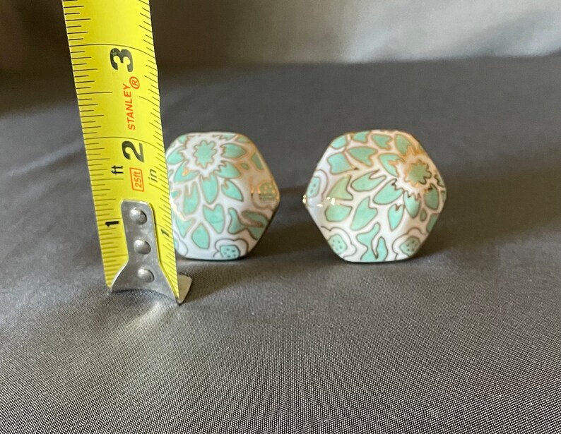 Pu&ograve; includere: Due pomelli esagonali in ceramica con base bianca, decorati con un motivo floreale verde acqua e dettagli dorati. Ogni pomello misura circa 2,5 cm di altezza. Questi pomelli decorativi sono ideali per mobili.