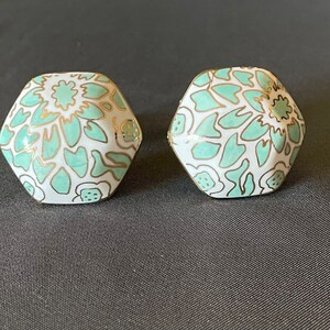 Pu&ograve; includere: Due orecchini esagonali con base bianca, decorati con un motivo floreale verde acqua e dettagli dorati. Gli orecchini sono presentati su uno sfondo grigio scuro, evidenziando il loro design e contrasto cromatico.