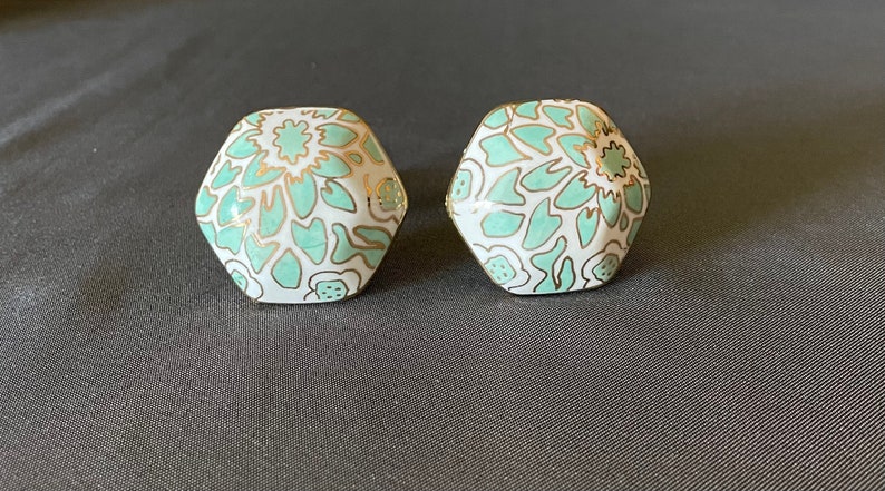 Pu&ograve; includere: Due orecchini esagonali con base bianca, decorati con un motivo floreale verde acqua e dettagli dorati. Gli orecchini sono presentati su uno sfondo grigio scuro, evidenziando il loro design e contrasto cromatico.