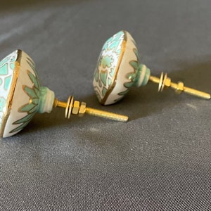 Pu&ograve; includere: Due pomelli decorativi per mobili. Presentano una base bianca ornata da un motivo floreale turchese e bordi dorati. Hanno ferramenta in ottone, tra cui un'asta filettata, rondelle e dadi. Per mobili.