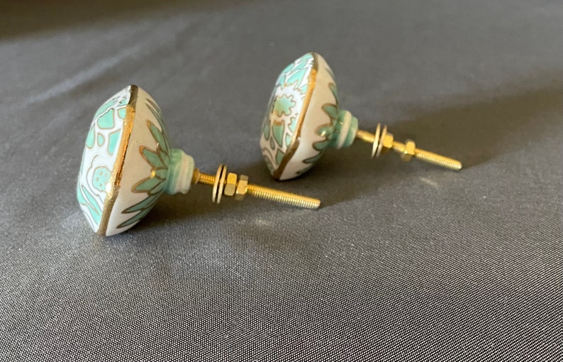 Pu&ograve; includere: Due pomelli decorativi per mobili. Presentano una base bianca ornata da un motivo floreale turchese e bordi dorati. Hanno ferramenta in ottone, tra cui un'asta filettata, rondelle e dadi. Per mobili.