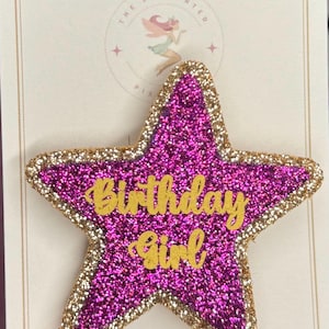 May include: A magenta glitter star-shaped decoration with gold glitter edging, featuring the words "Birthday Girl" in gold script. The background is a cream-coloured card.