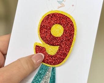 May include: Glittery number nine birthday decoration. The number is red with yellow accents and a teal, silver, and white streamer. The decoration is on a white card with a logo that says "The Enchanted".