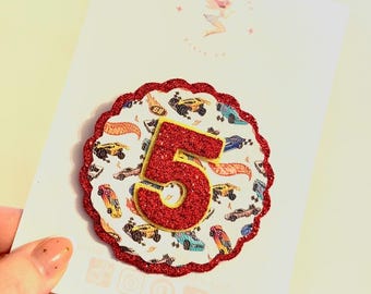 May include: A handmade birthday card featuring a red glitter number 5. The number is placed on a white background with a red scalloped border. The background has a pattern of colorful toy cars.