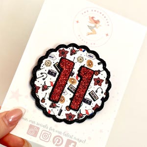 May include: A circular scalloped badge featuring a white background with a repeating pattern of small icons including cameras, cassettes, and flowers. The number 11 is prominently displayed in the centre in large, textured, glittery red font.