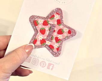 May include: Two star-shaped hair clips. The inner star features a pink background with a pattern of red strawberries and white flowers. The outer star has a glittery pink border. The card reads "The Enchanted Pixie Co."