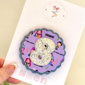 May include: A purple and glittery number eight-shaped pin. The pin has a scalloped edge with a glittery border. The center of the pin is a large number eight covered in silver glitter. The pin has a purple background with cartoon characters and the text "HUNTRIX".