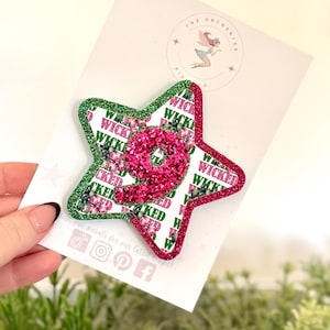 May include: A star-shaped badge with a green and red glitter border. The badge has the word "WICKED" repeated over a floral pattern and a glittery pink number "9". The badge is held over a white card with a logo.