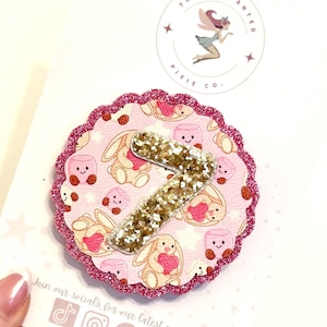 May include: A circular scalloped badge featuring a pink pattern of cartoon bunnies and marshmallows with a large gold glitter number seven centred on top. The badge has a bright pink glitter border and is attached to a white display card.