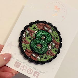 May include: A circular badge with a green glitter number 8. The badge has a black scalloped edge and a patterned background. The badge is on a white card with social media icons and the text 'Join our socials'.