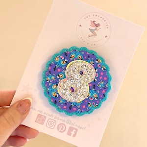 May include: A glittery number eight, with a silver glitter finish, is centered on a purple and teal circular design. The design features a repeating cartoon cat face pattern. The item is on a white card with the text "The Enchanted Pixie Co."